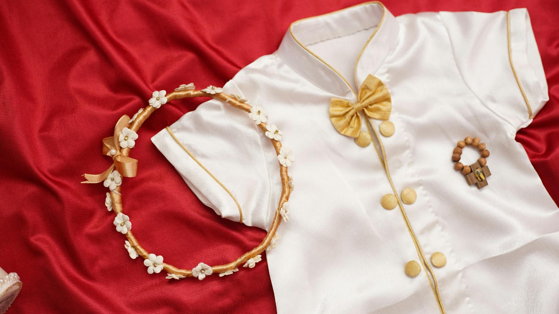 Białe ubranko do chrztu z wiankiem i różańcem na czerwonym tle